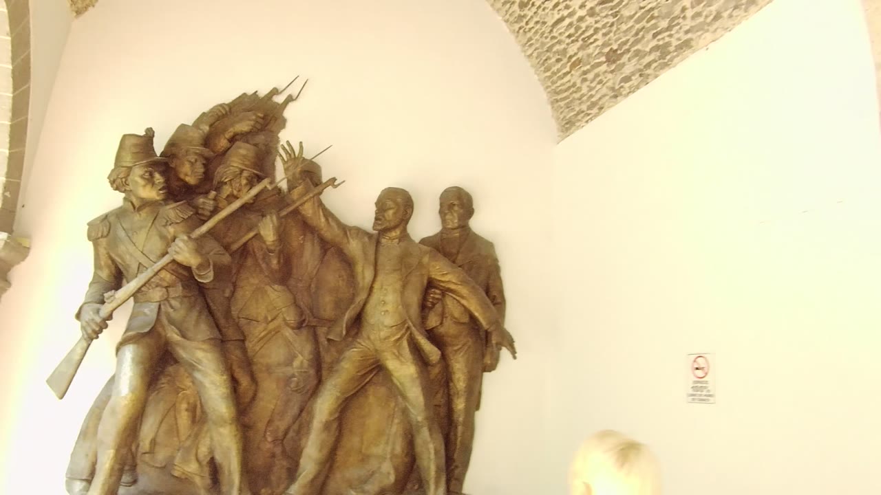Wide upper view of a bronze historical relief sculpture in Guadalajara, Mexico, framed beneath stone arches with a pale wall behind it and no visible people.