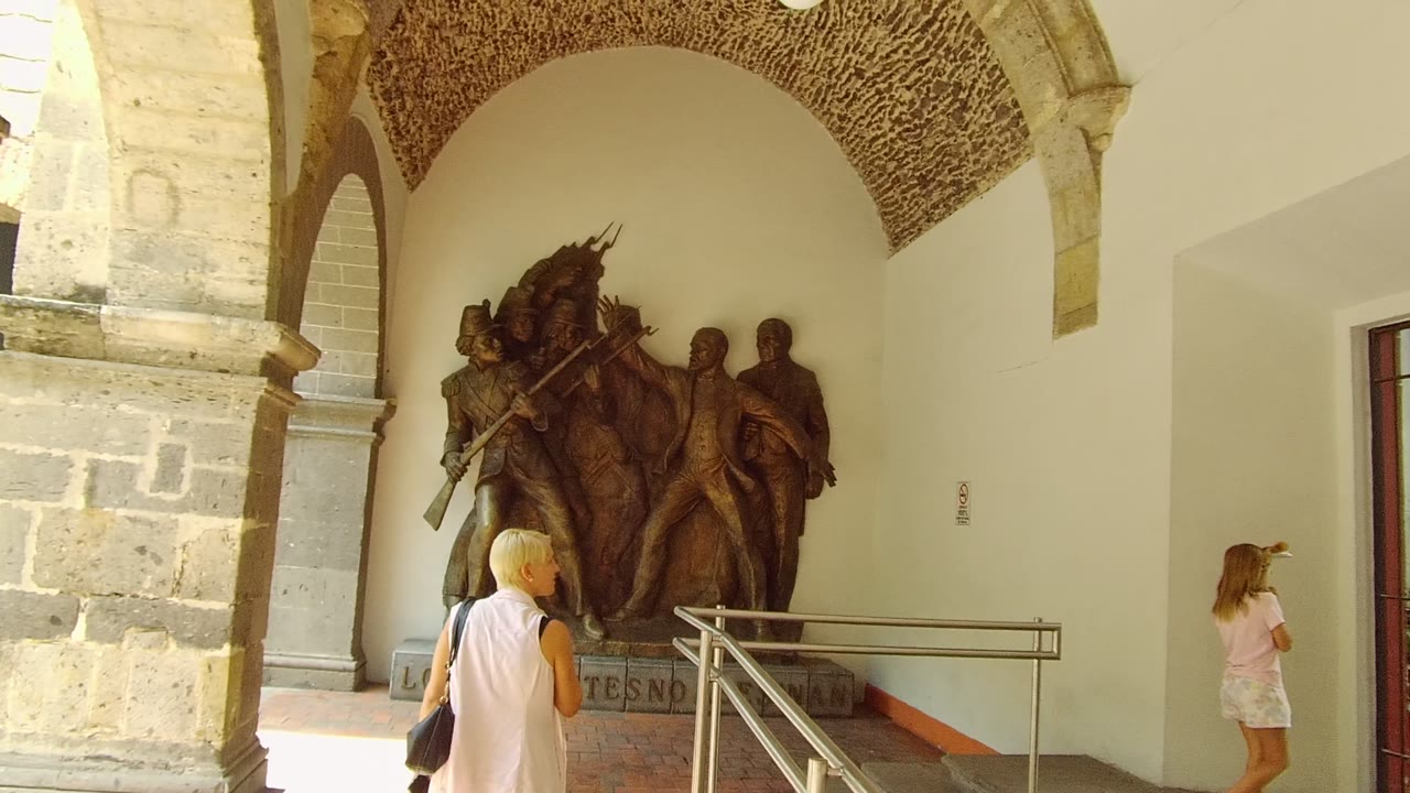 A woman stands near a bronze historical relief beneath stone arches in a Guadalajara corridor, with railings and warm indoor light visible.