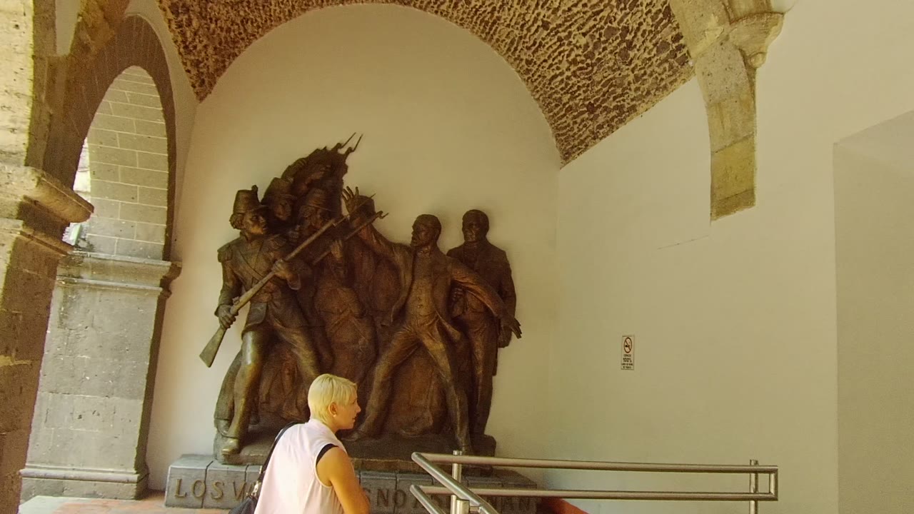 The woman moves closer to the bronze relief in the Guadalajara corridor, passing in front of the wall railing beneath the arch.