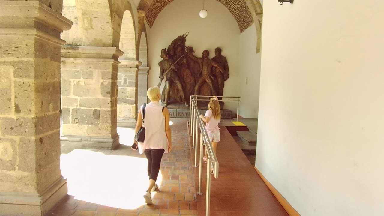 A woman and child walk away from the camera under a stone archway in Guadalajara, Mexico, with brick flooring, pale walls, metal railings, and a large statue ahead.