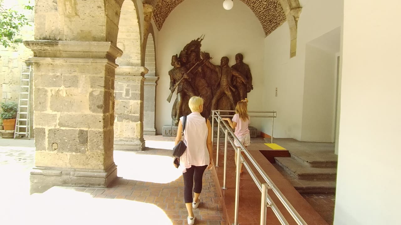 The pair continues through the Guadalajara stone arcade, passing a bright opening on the left while the statue, railing, and white-walled alcove remain ahead.