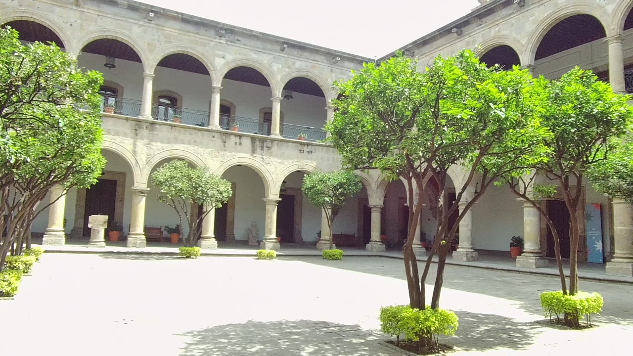 Wide view of a Guadalajara stone courtyard with two levels of arches, leafy trees near the center, bright paving, and no visible people.