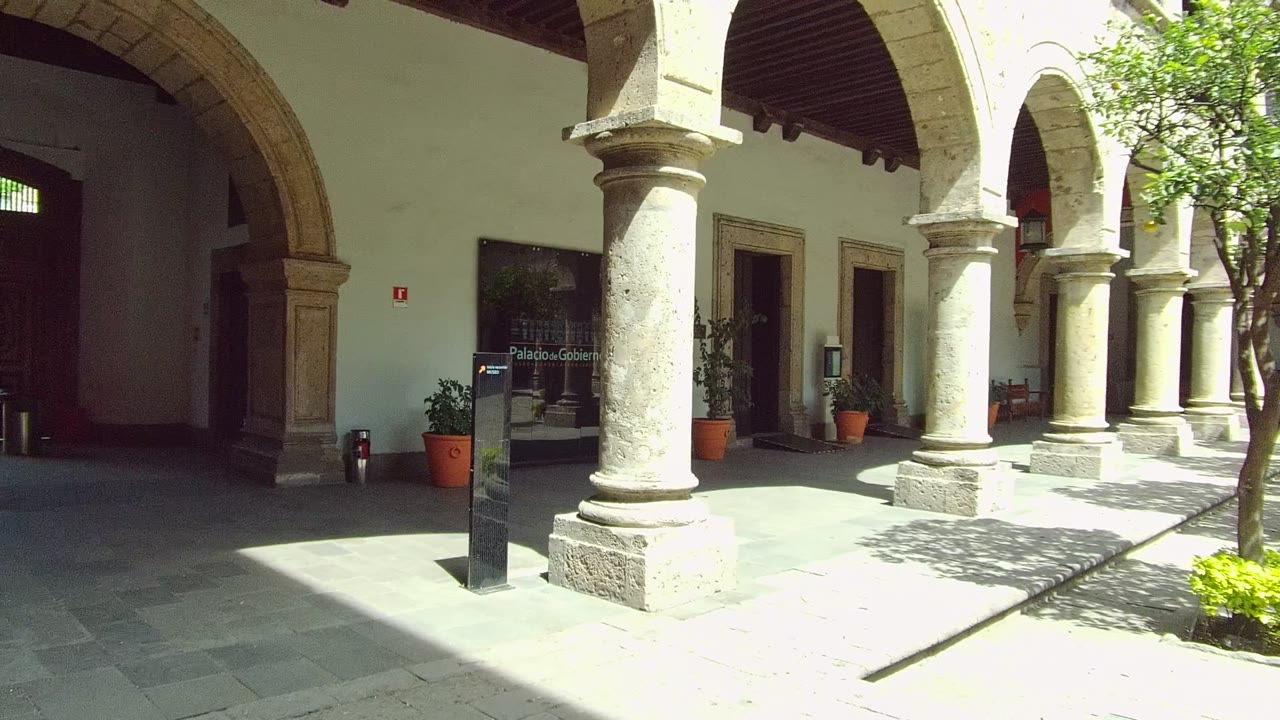 A Guadalajara stone courtyard shows pale columns, arched walkways, potted plants, a freestanding sign, a small tree, sunlit paving, and no visible people.