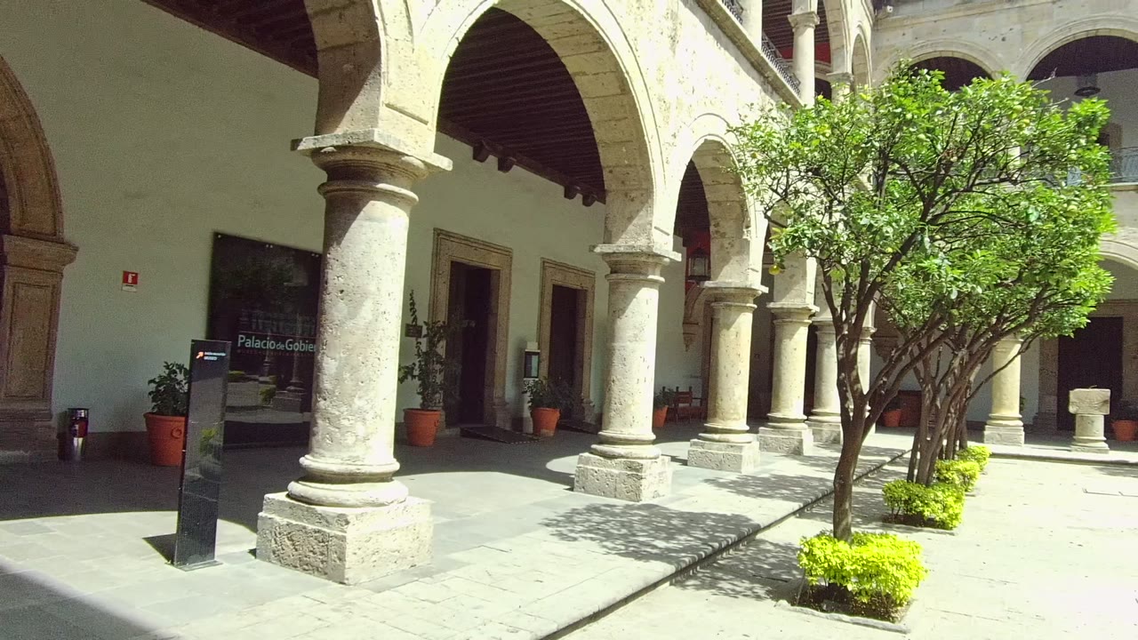 The Guadalajara courtyard view moves farther along the arcade, showing thick stone columns, leafy trees, planted beds, shaded doorways, sunlit paving, and no visible people.