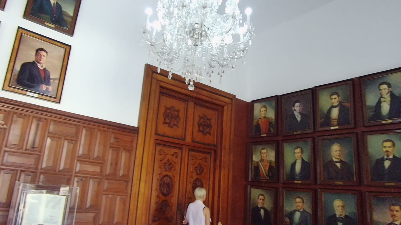 At 0:00 in Guadalajara, Mexico, a bright paneled interior shows a chandelier, carved wooden door, framed historical portraits, and a person in a white sleeveless shirt near the lower center.