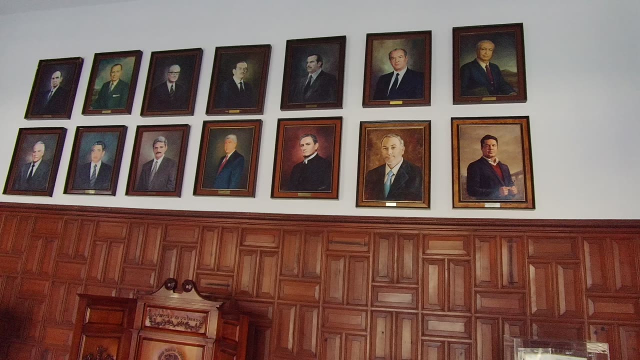 Rows of framed portraits hang high on a white wall above warm wooden paneling in a Guadalajara interior, with no visible people in this opening frame.