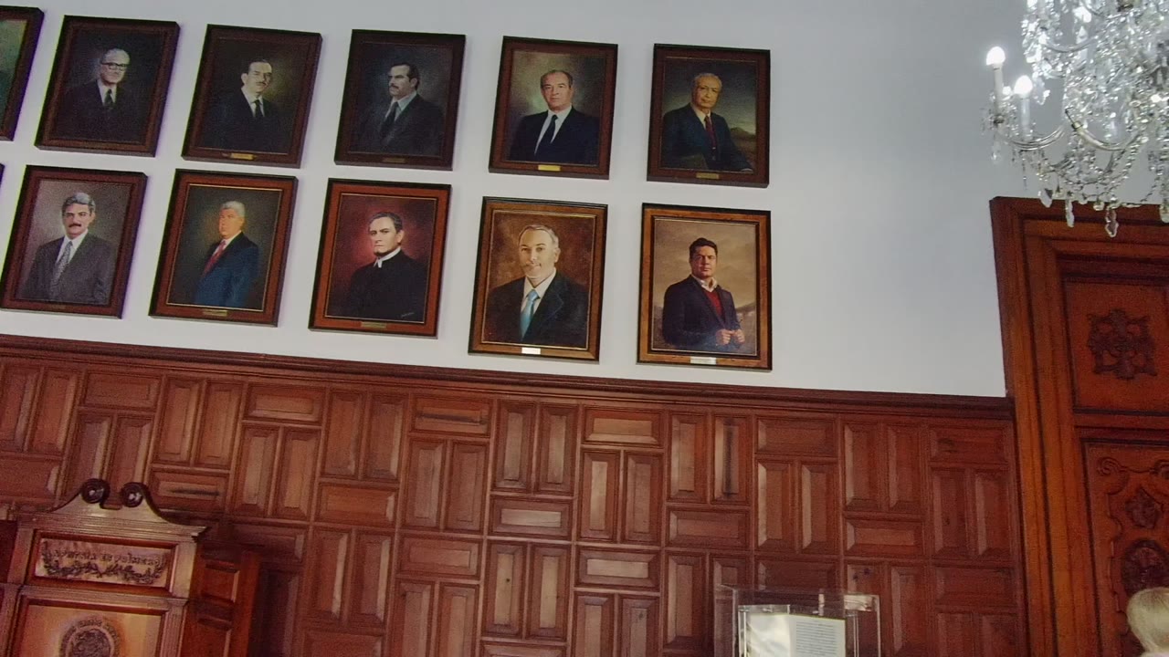 A crystal chandelier begins to enter at the top right while framed portraits and carved wooden wall panels remain the main view in the Guadalajara room, with no visible people.