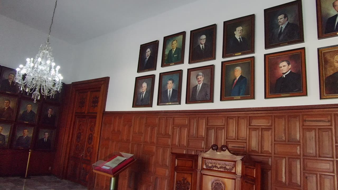 Guadalajara, Mexico interior with a chandelier at left, framed portraits above wooden wainscoting, a red book on a podium, and no visible people.
