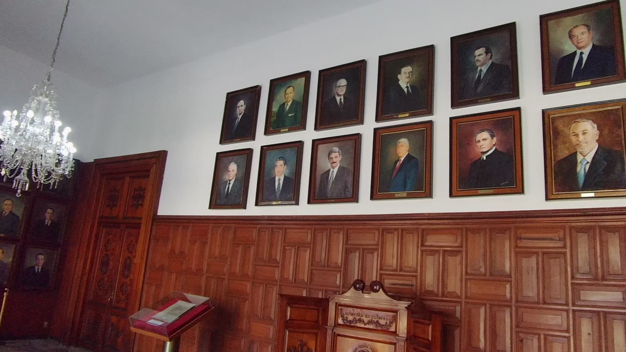 Guadalajara, Mexico room with rows of framed portraits over carved wooden wainscoting, a chandelier near the right edge, and no visible people.
