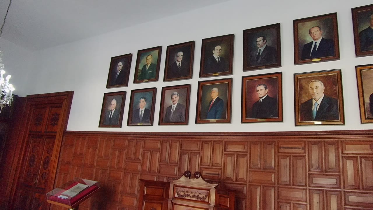 Guadalajara, Mexico interior wall with framed portraits in two rows, warm wood wainscoting, a red book stand at lower left, and no visible people.