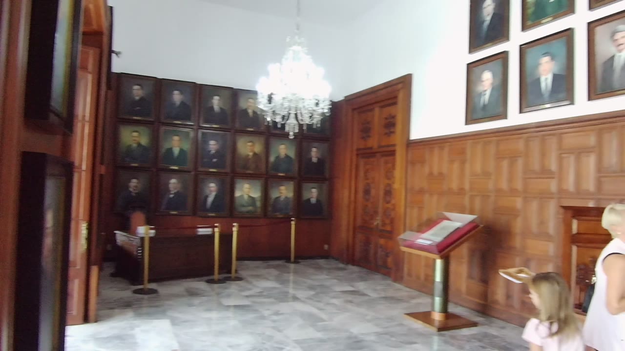 Wide view of a Guadalajara portrait room with wood paneling, a chandelier, display stand, and a woman with a child partially visible at the right edge.