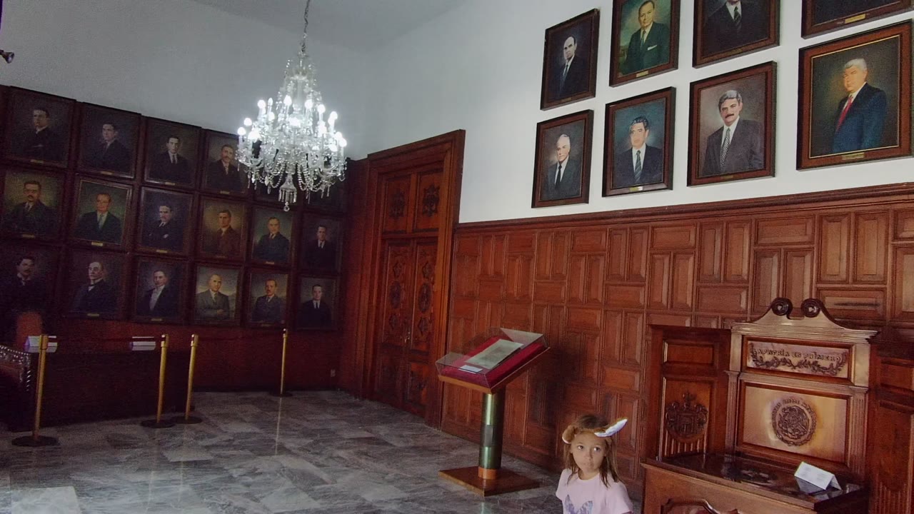 Wide Guadalajara room view with framed historical portraits above wood paneling, a chandelier overhead, a display stand, and a child visible near the right foreground.