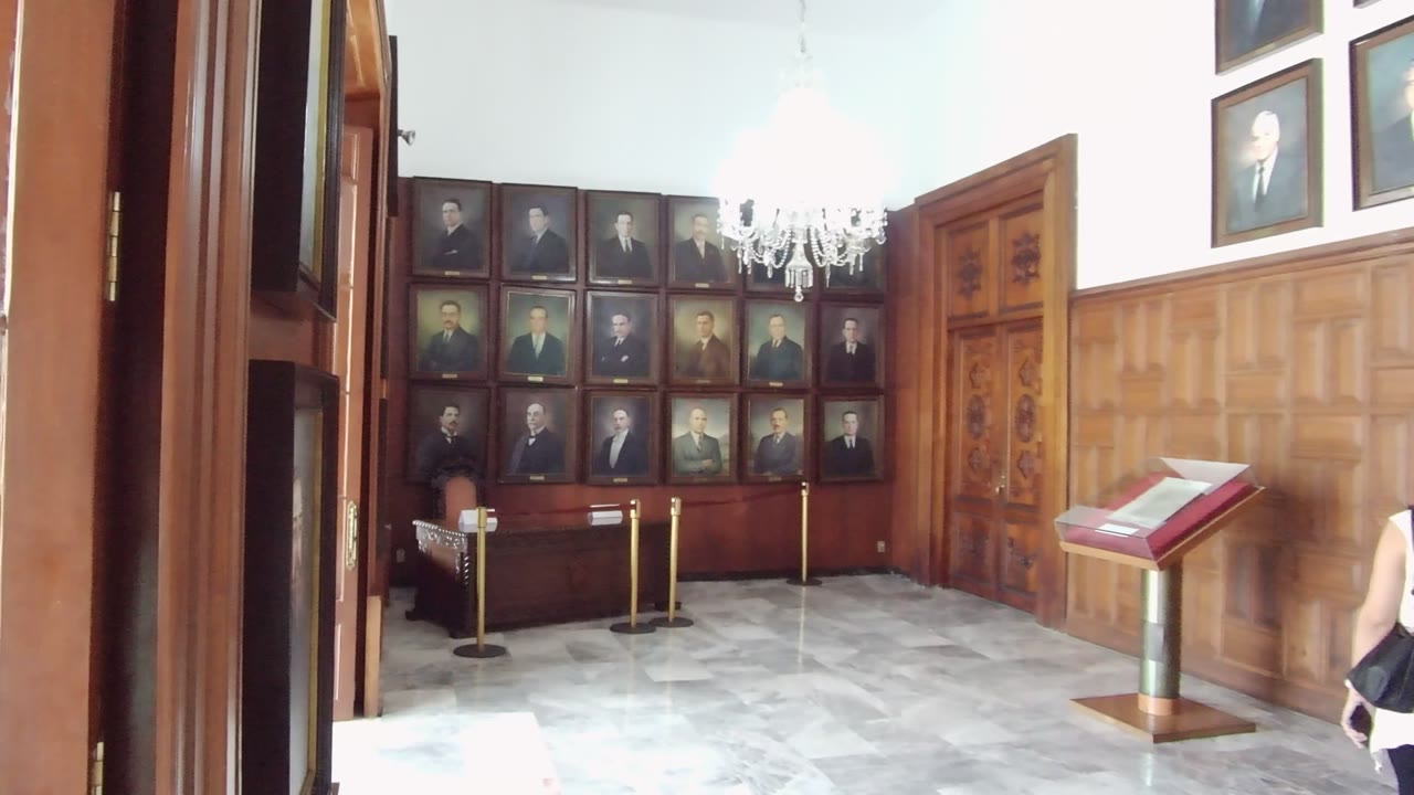 A Guadalajara museum-like interior shows wood paneling, framed historical portraits, a chandelier, marble floor, rope barriers, and a red display stand, with a partial visitor at the right edge.