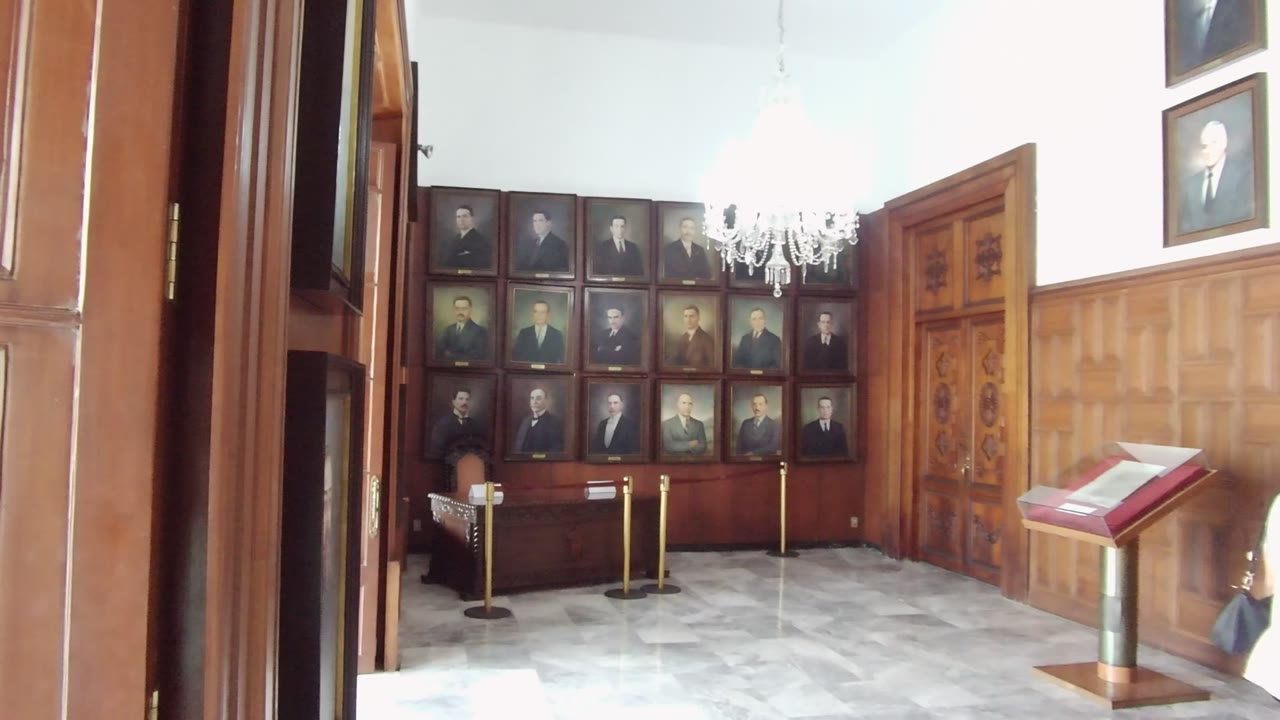 A doorway view into the Guadalajara room shows dark foreground trim, paneled walls, rows of framed portraits, a bright chandelier, marble flooring, rope barriers, and the angled display stand.