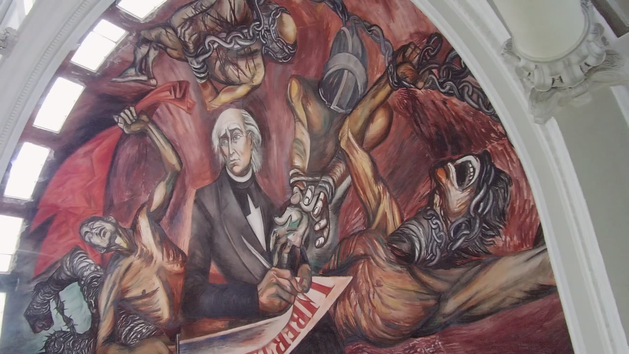 Opening frame of a Guadalajara interior mural showing a historical figure writing beside chained painted figures, with bright arched window light at left and no visible people.