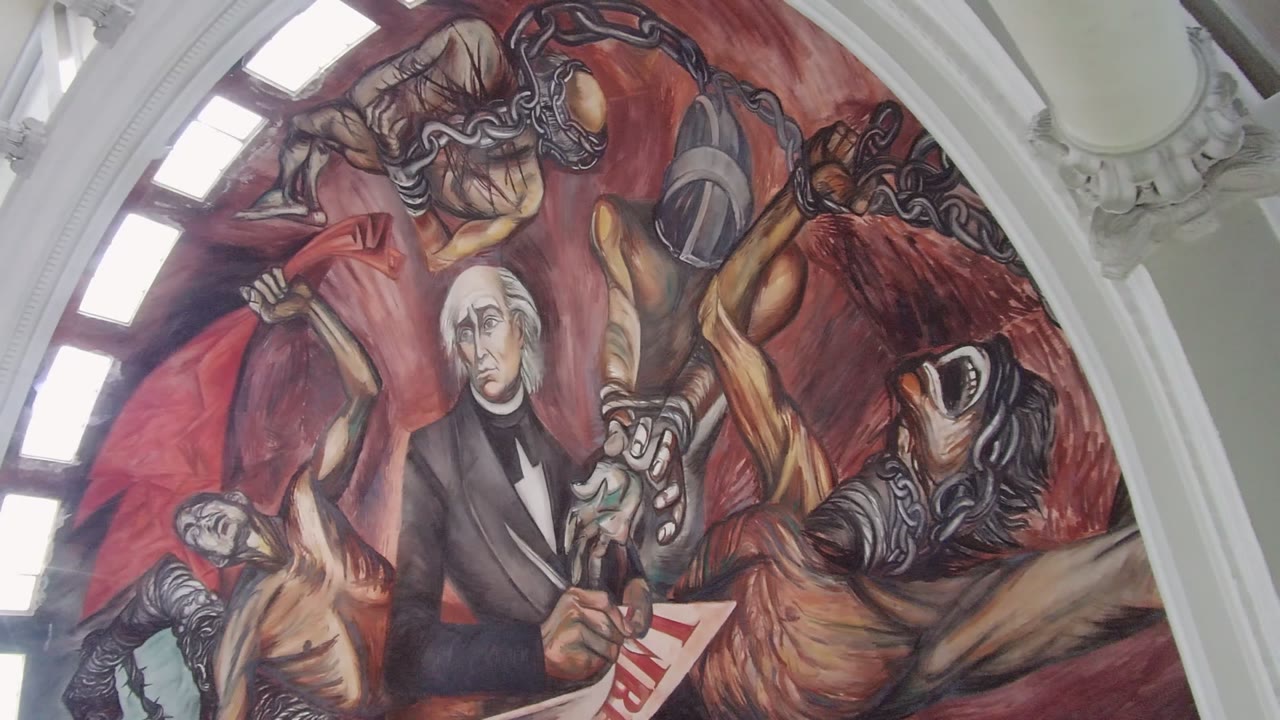 At 0:02, the frame shows the Guadalajara wall mural under a white interior arch, with the red painted background, chains, and liberty document visible, and no visible people.
