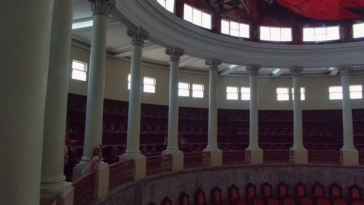 Opening frame inside a domed building in Guadalajara, Mexico, showing white columns around a circular balcony, bright upper windows, the edge of a red ceiling mural, and small people near the balcony.