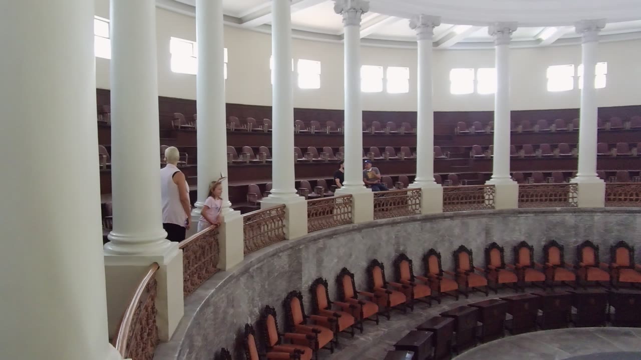 Guadalajara hall interior at the start of the clip, showing white columns, upper windows, curved balcony railing, brown seating below, and people at the left side.