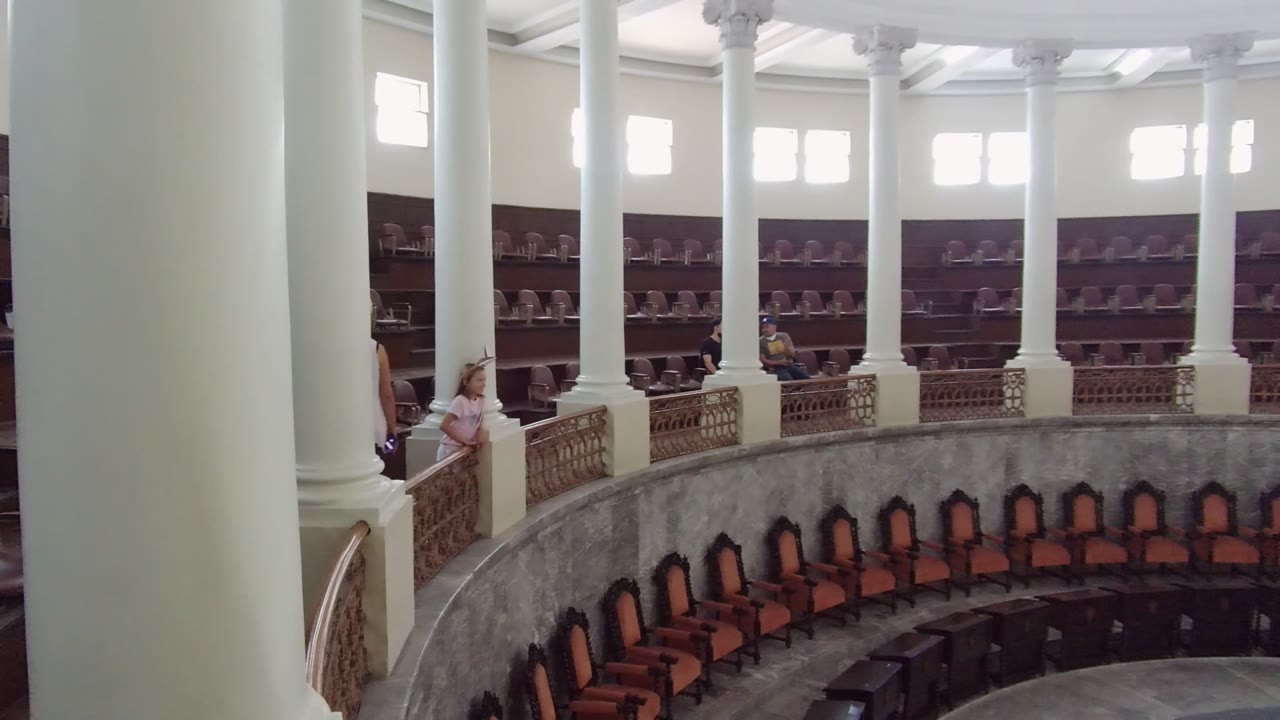 Grand interior balcony with white columns, high windows, rows of chairs, and people by the railing.