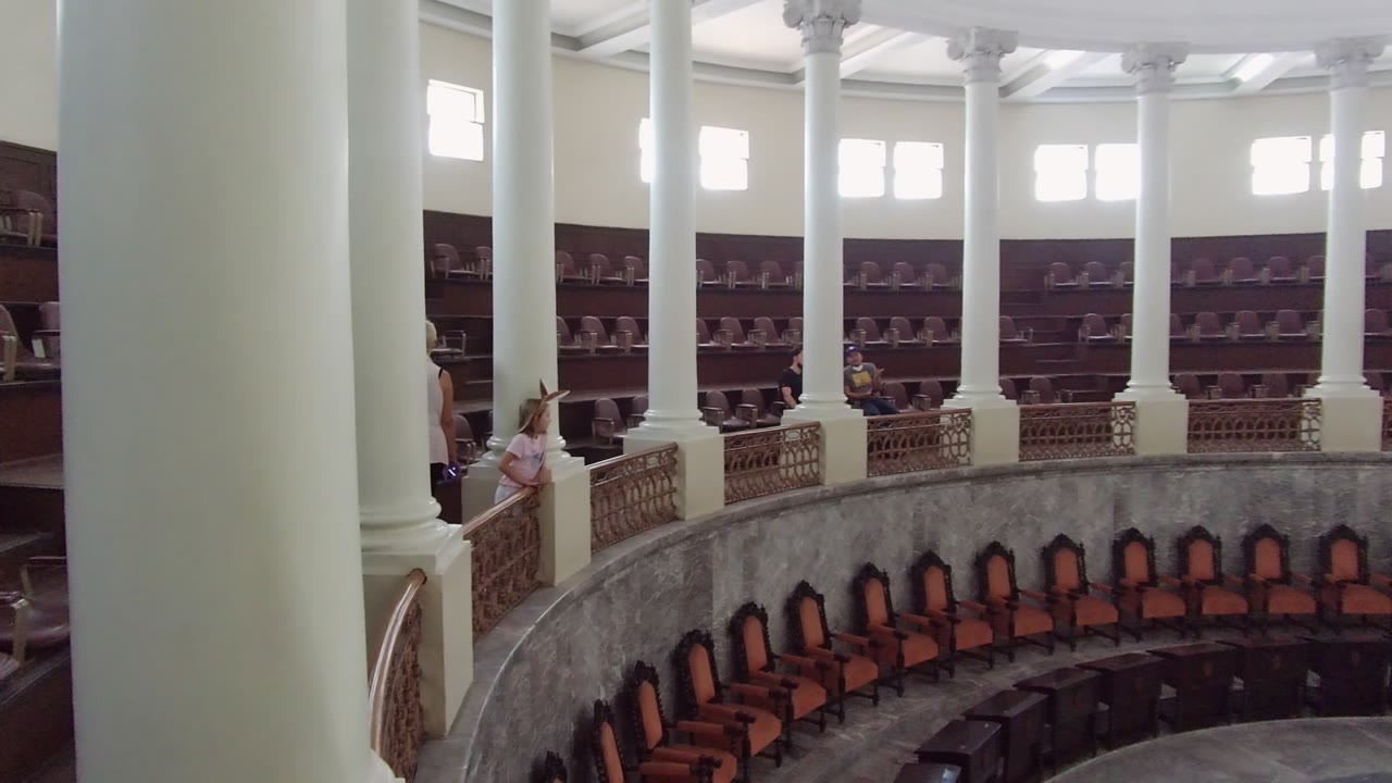 Guadalajara hall interior at two seconds, with the balcony and people shifted slightly left in the frame while columns and brown seating remain prominent.