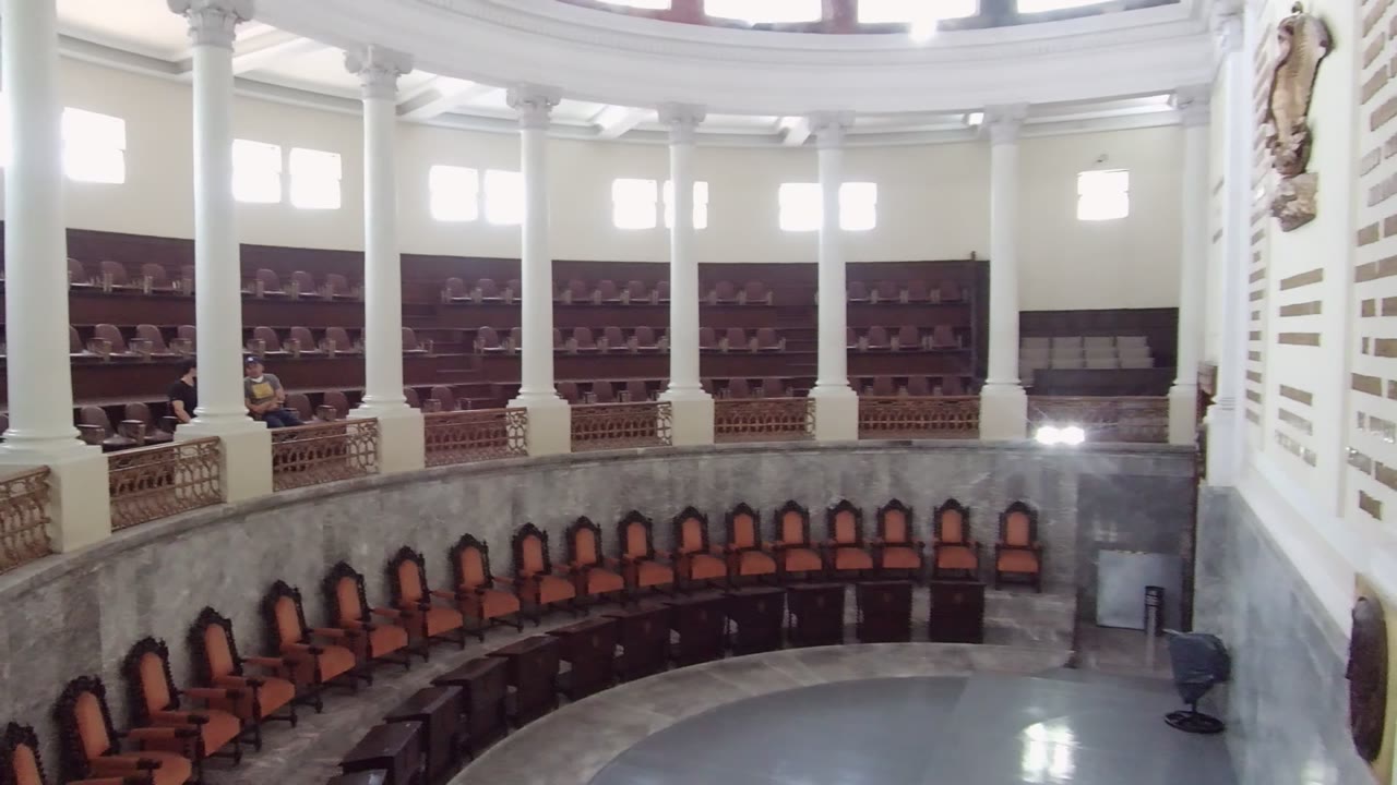 Wide view of a circular interior in Guadalajara, Mexico, with white columns, bright upper windows, curved railings, orange-cushioned wooden chairs, and no visible people in the main room.