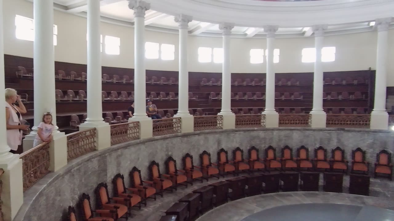 The adult and child are clearer at the left side of the balcony in the Guadalajara interior, with white columns, decorative railing, and empty orange seats across the circular room.