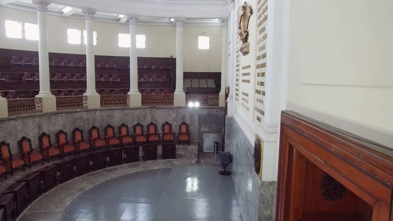 Elevated view of a Guadalajara assembly hall with curved orange chairs, dark desks, white columns, bright high windows, and no visible people.