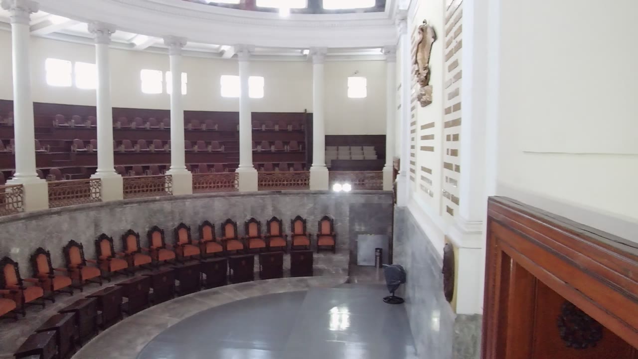 Elevated side view across the Guadalajara assembly hall, showing white columns, bright upper windows, rows of orange seats, dark desks, and no visible people.