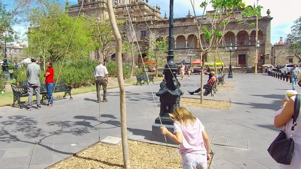 Opening frame of a Guadalajara public square with a black lamppost near center, pedestrians at left, benches, young trees, and a historic arched building in the background.
