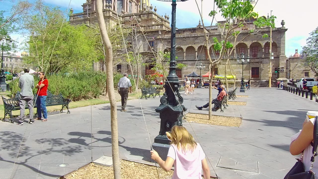 At two seconds, a child in a pink shirt is low in the foreground beside hanging ropes, while pedestrians and seated people remain visible before the Guadalajara historic building.