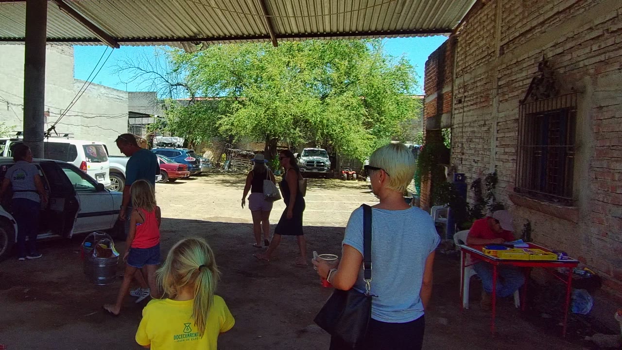 Adults and children stand and walk under a shaded carport in Tequila, Jalisco, with a brick wall, parked vehicles, and a sunlit courtyard beyond.
