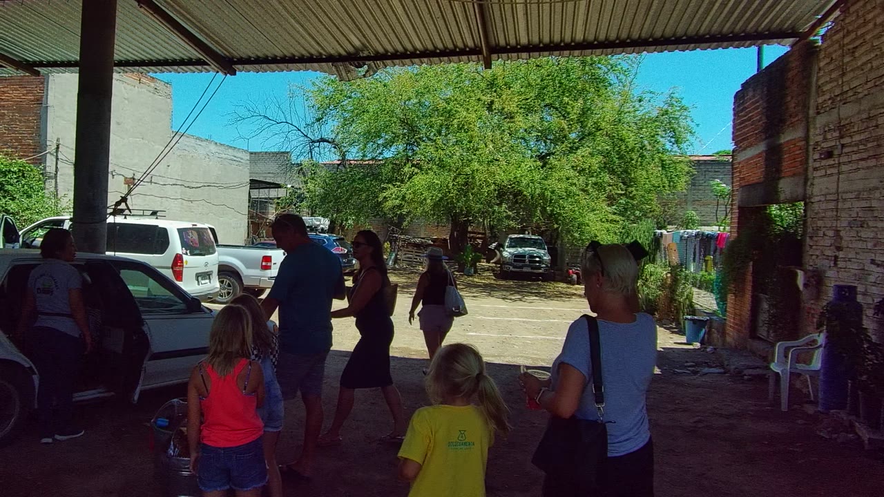 Adults appear to talk near children under the carport in Tequila, Jalisco, with a brick wall on one side, parked cars, laundry, and a leafy tree in the bright background.