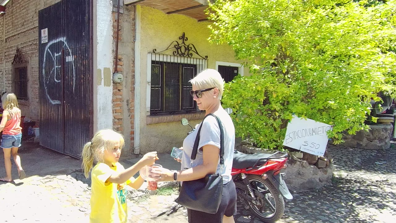 In Tequila, Jalisco, a woman in sunglasses and a young girl in a yellow shirt stand near a brick wall, dark metal gate with graffiti, leafy tree, and red motorcycle.