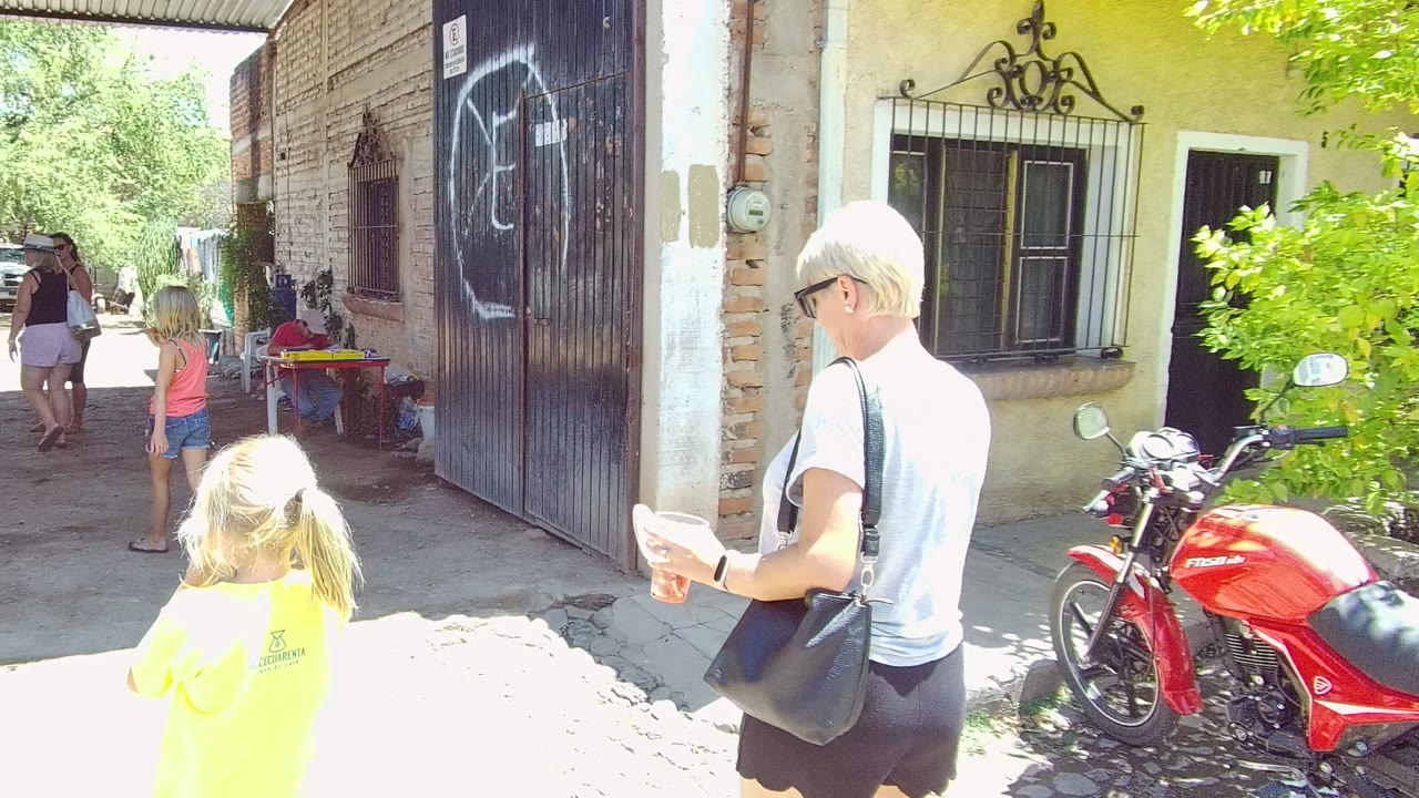 In Tequila, Jalisco, the woman holds a cup while the child walks ahead near a red motorcycle, a dark graffitied gate, barred windows, and background pedestrians.