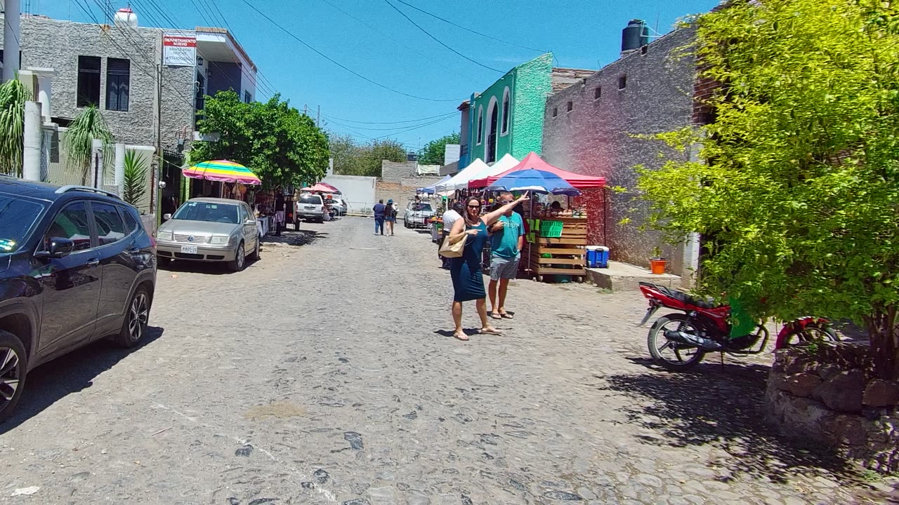 Two adults walk toward the camera on a cobblestone street in Tequila, Jalisco, with parked cars, a red motorcycle, stone buildings, and colorful market umbrellas visible.