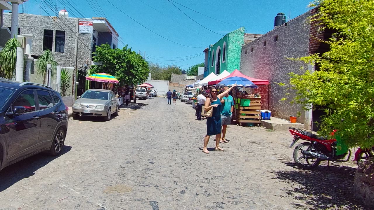 People walking near market stalls on a cobblestone lane with cars and umbrellas.