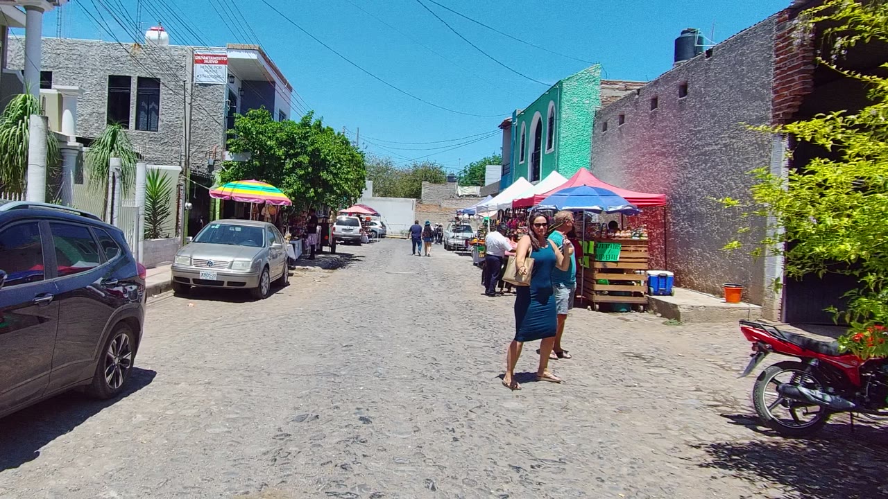 A woman and man move closer on the sunlit street in Tequila, Jalisco, with a red motorcycle in the right foreground and vendor umbrellas beyond them.