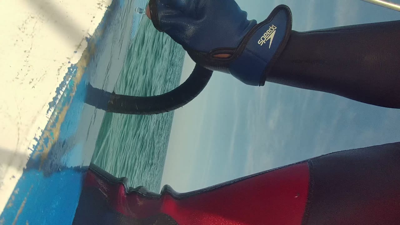 Cabo Pulmo water activity frame with the boat edge, green ocean, cloudy sky, and a partial wetsuit-clad person wearing a blue glove near a black handle.
