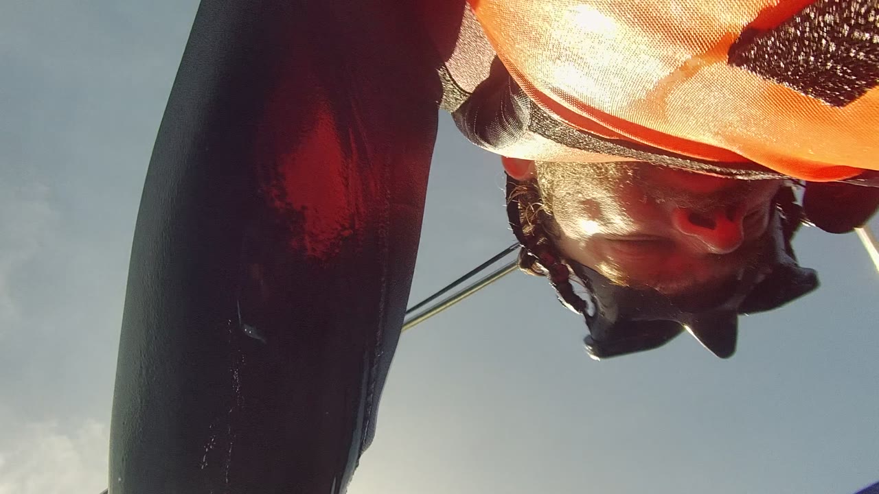 An upside-down view shows the swimmer's partial face, dark wetsuit sleeve, and orange life vest above the camera in Cabo Pulmo, with bright sky behind them.