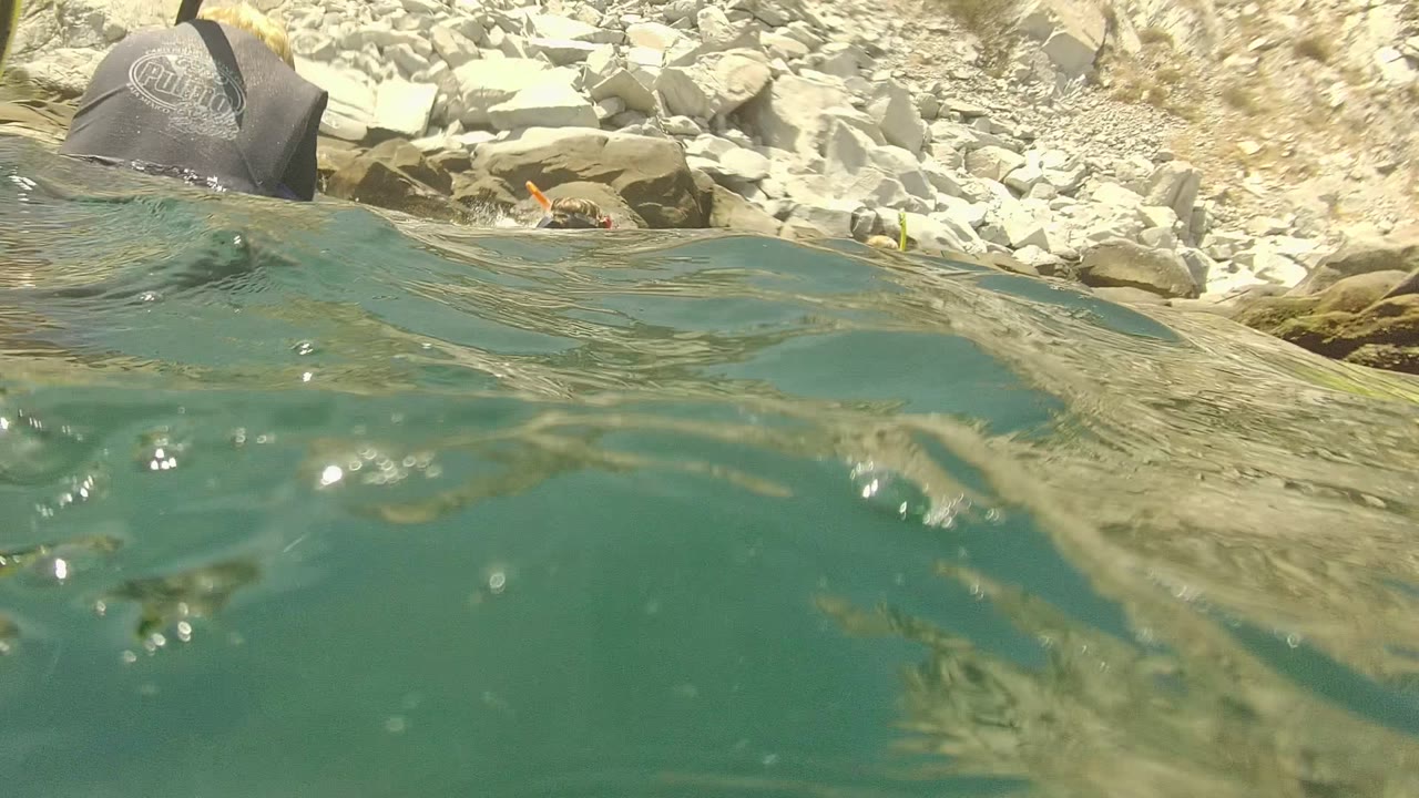 Waterline frame in Cabo Pulmo, Mexico showing turquoise water, rocky shoreline, glare on ripples, and snorkel gear partly visible near the surface with no recognizable faces.