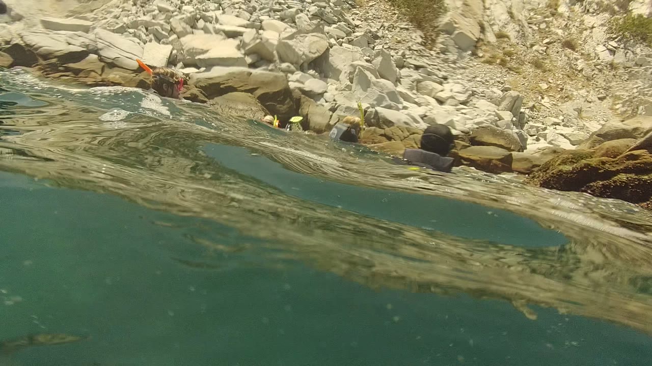 Waterline frame in Cabo Pulmo, Mexico showing rocky shoreline above the rippled surface, partially submerged snorkelers, and yellow fins near the rocks with no recognizable faces.