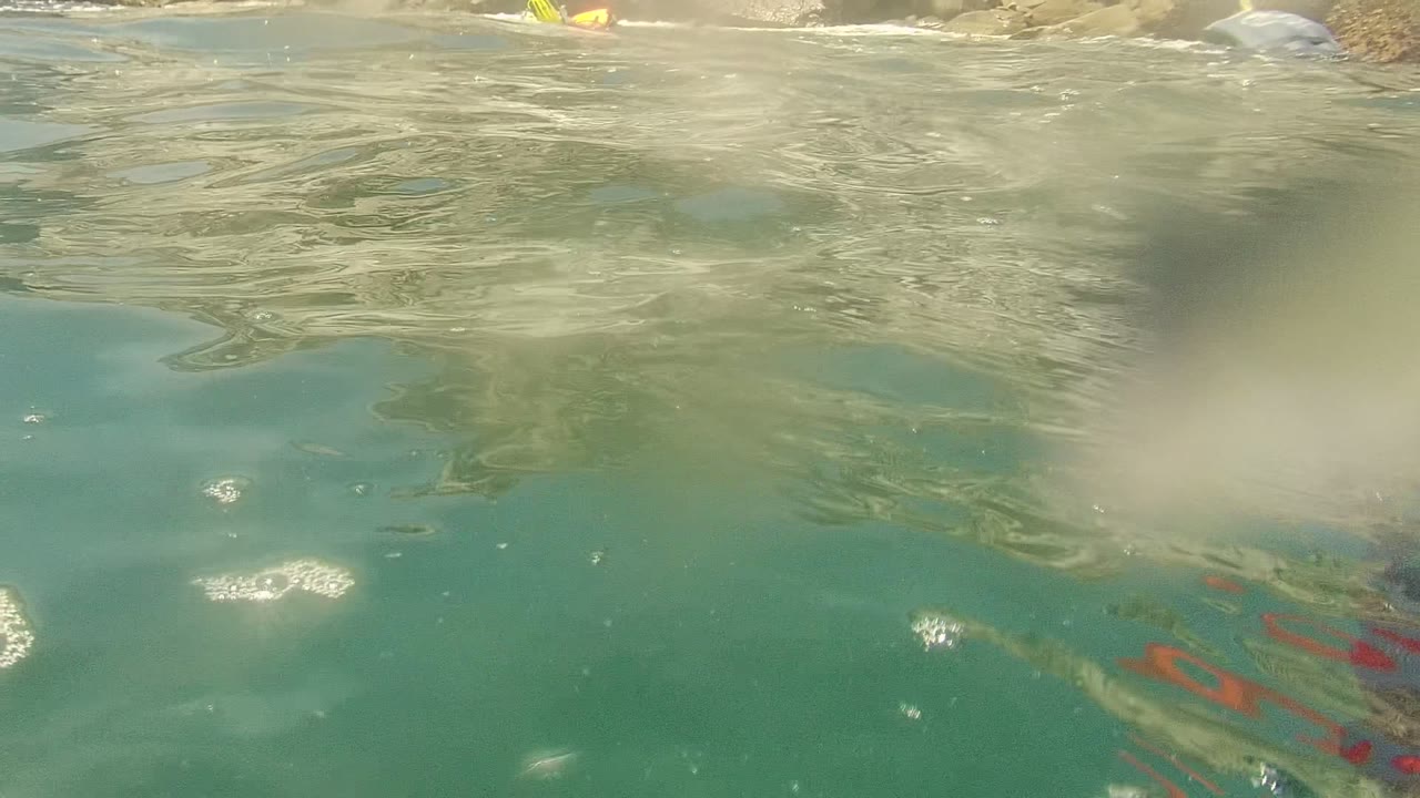 Water-level view in Cabo Pulmo, Mexico showing clear green-blue ocean with partial snorkel gear at the top edge and pale rocks beyond the rippled surface.