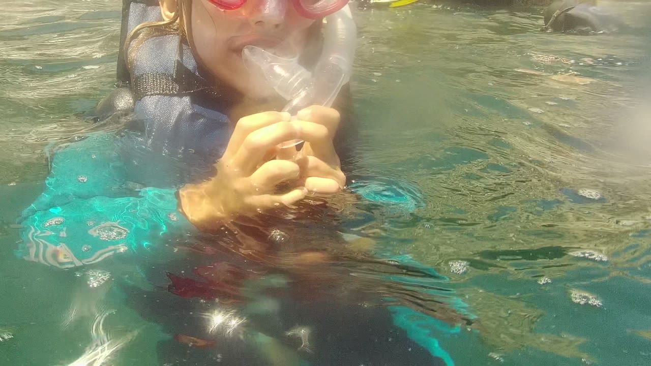 A snorkeler in Cabo Pulmo, Mexico wearing a pink mask and dark life vest holds the snorkel mouthpiece at the surface, with clear green-blue water and rocks behind.