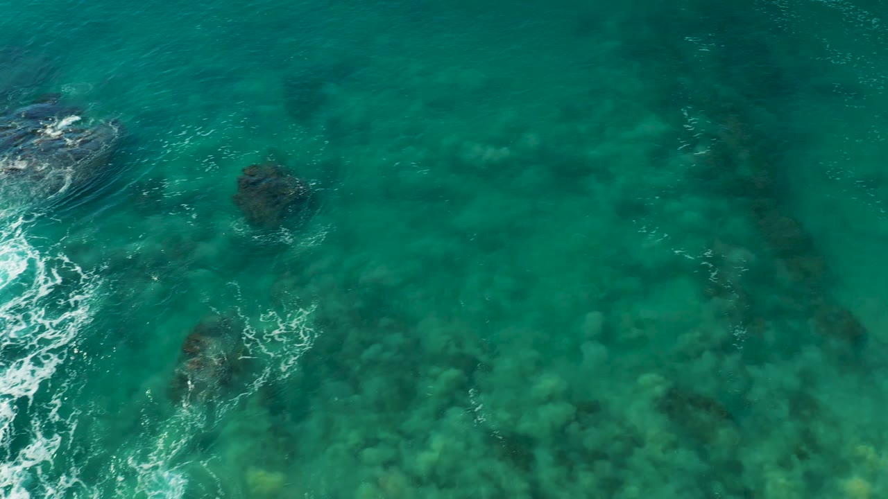 Overhead view of clear turquoise water at La Fortuna, Baja California Sur, with submerged rocks, darker seabed patches, white foam along the left edge, and no visible people.
