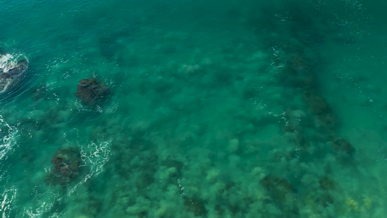 Clear green-blue water over seabed rocks with a small white wave break near center.