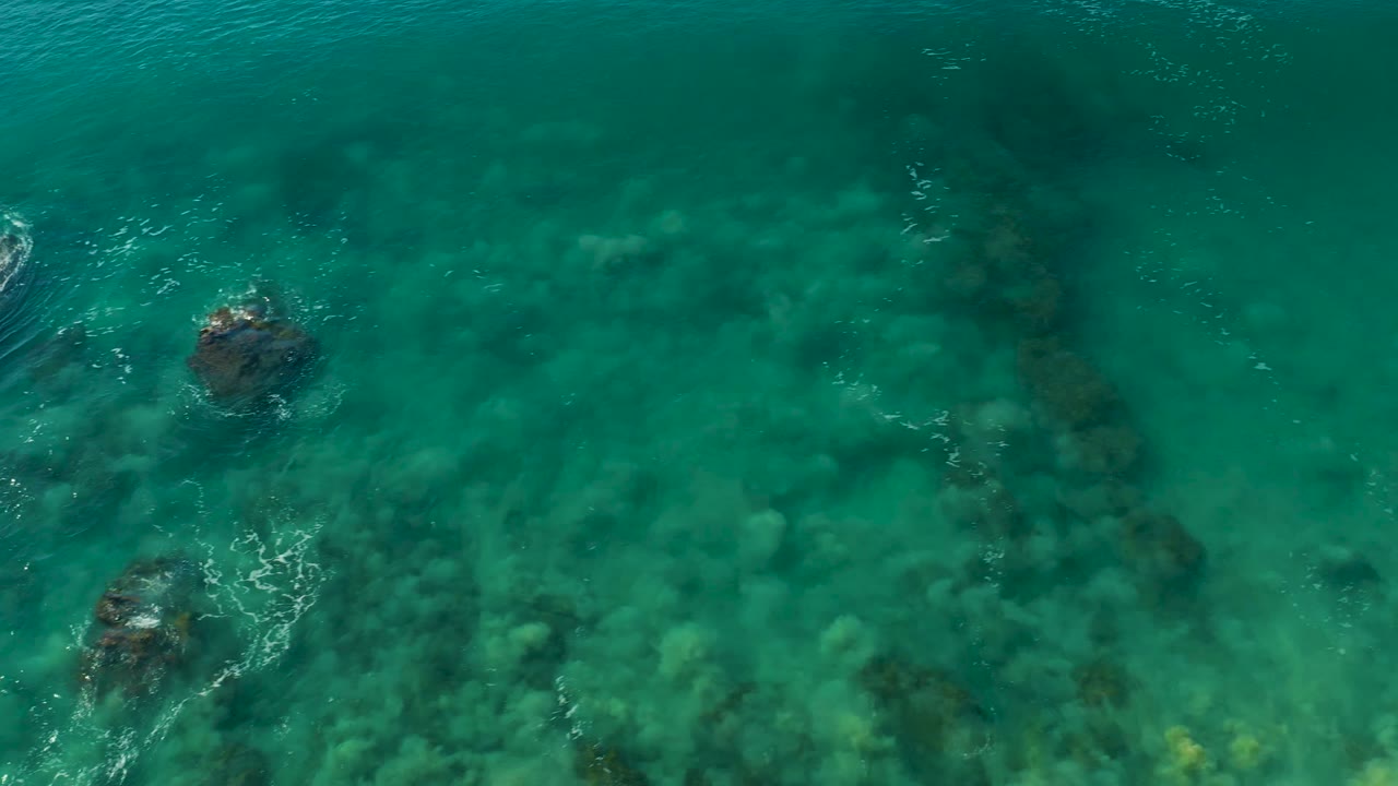 Top-down La Fortuna ocean frame with turquoise water, dark submerged rocks, faint coral-like seabed texture, and thin whitewater streaks over the rocks, with no visible people.