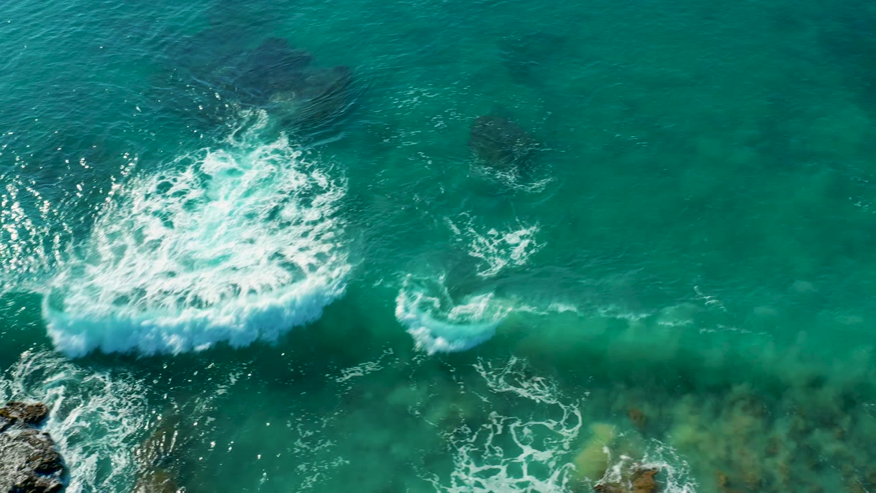 Aerial view of La Fortuna coastal water with a dense white breaking wave patch above turquoise shallows and darker rocks below, with no visible people.