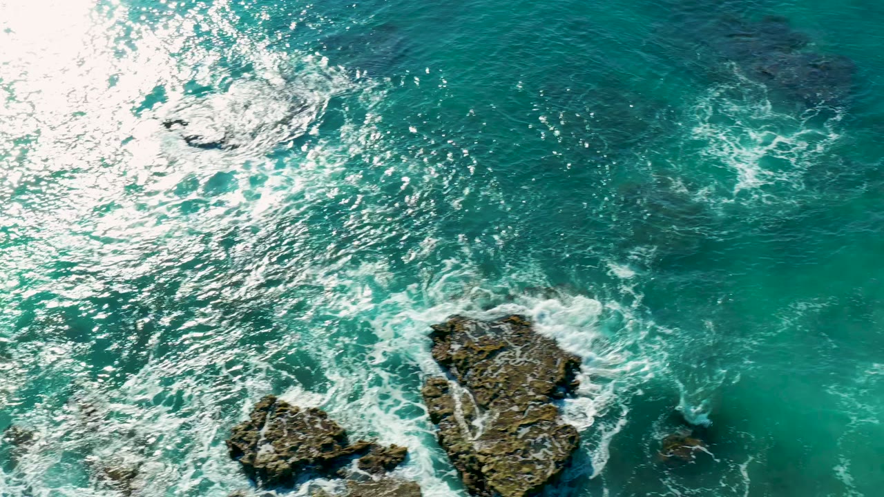 Top-down view of turquoise water and white foam surrounding rocky outcrops at La Fortuna, Baja California Sur, with no visible people.