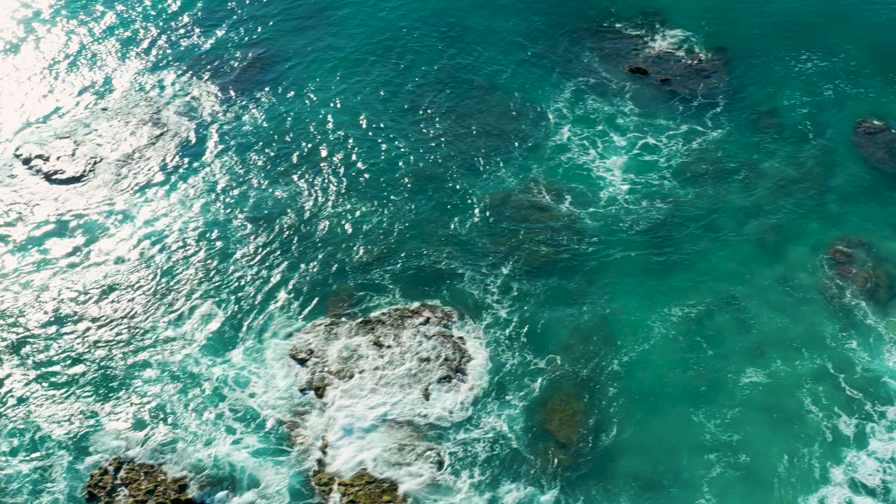 Turquoise sea viewed from above as white surf circles a central rock and smaller dark rocks at La Fortuna, Baja California Sur, with no visible people.