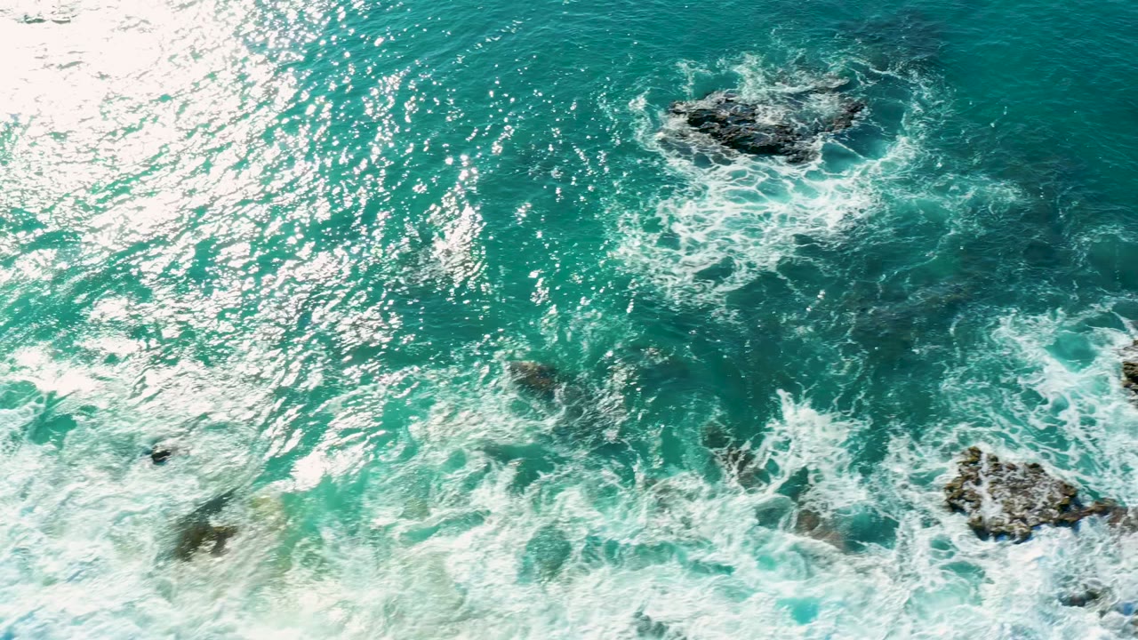 Overhead view of turquoise water and white foam forming around dark ocean rocks at La Fortuna, Baja California Sur, Mexico, with no visible people.